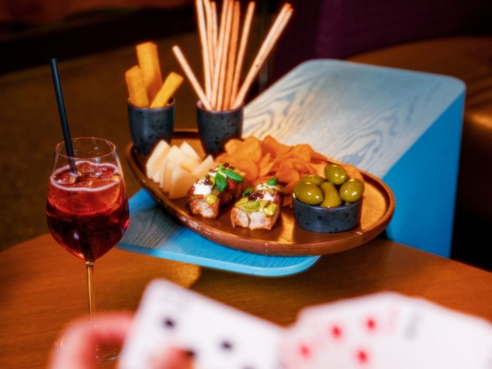 A table with an Spritz drink, a platter of olives, cheese, breadsticks, chips, and bruschetta. A hand holding playing cards is visible in the foreground.