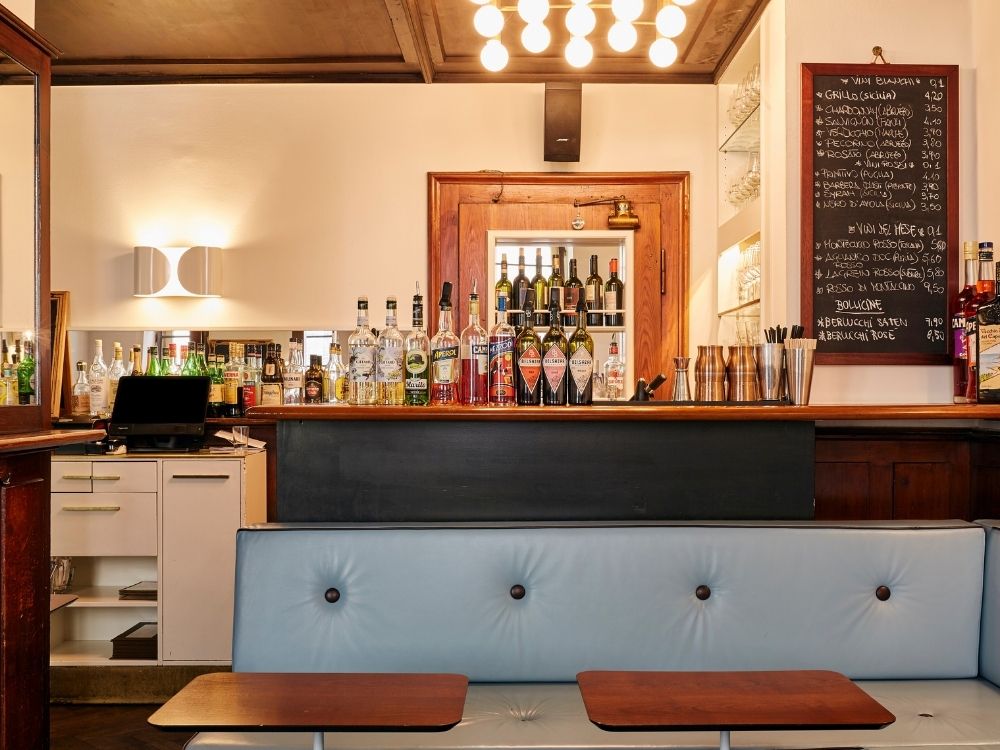Cozy bar interior with a variety of liquor bottles and wine displayed on a wooden counter. A chalkboard menu is on the right. Warm lighting creates an inviting atmosphere.