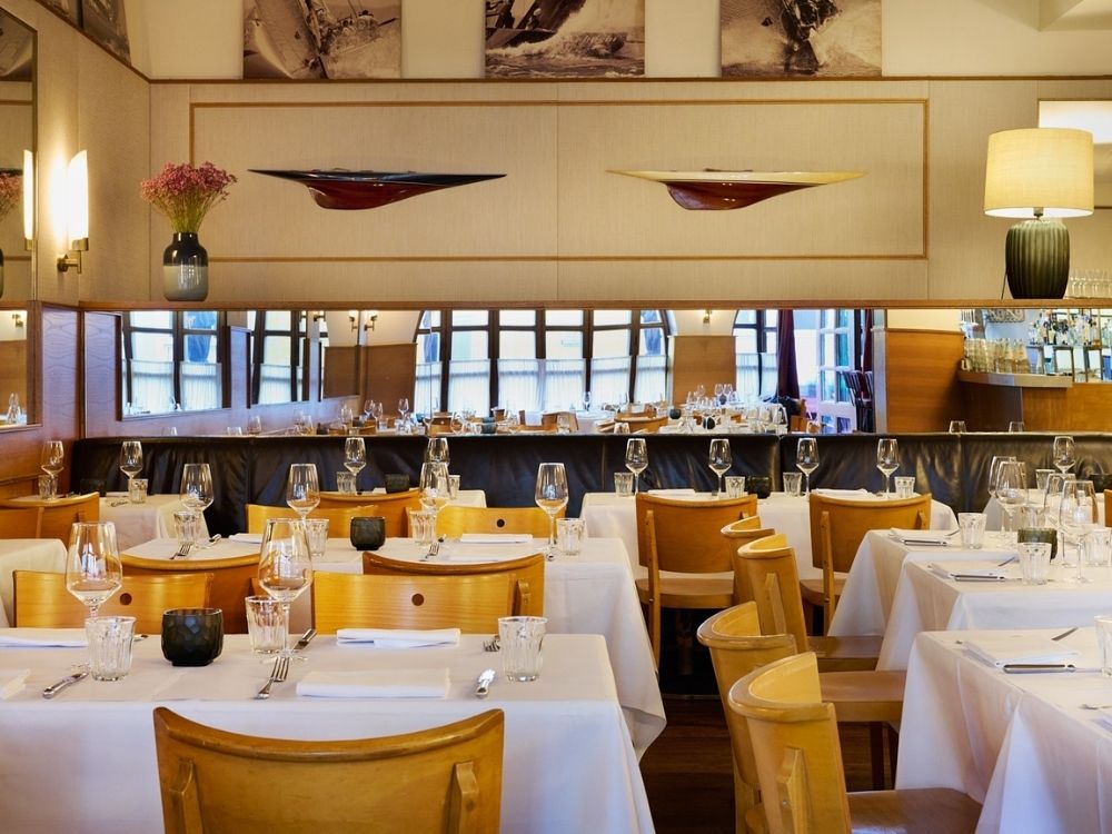 Elegant restaurant interior with neatly arranged tables, white tablecloths, wooden chairs, and carefully set glassware. Warm lighting and wall art create a cozy ambiance.