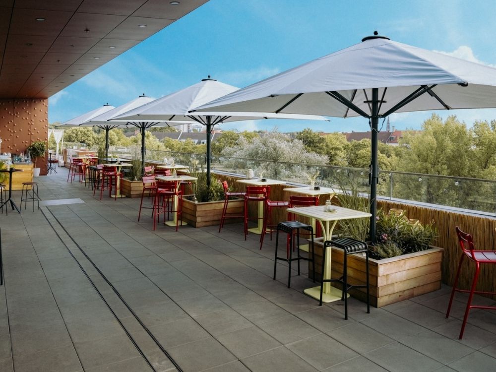 Outdoor dining area with white umbrellas, colorful tables, and red chairs overlooking lush greenery and a clear blue sky.