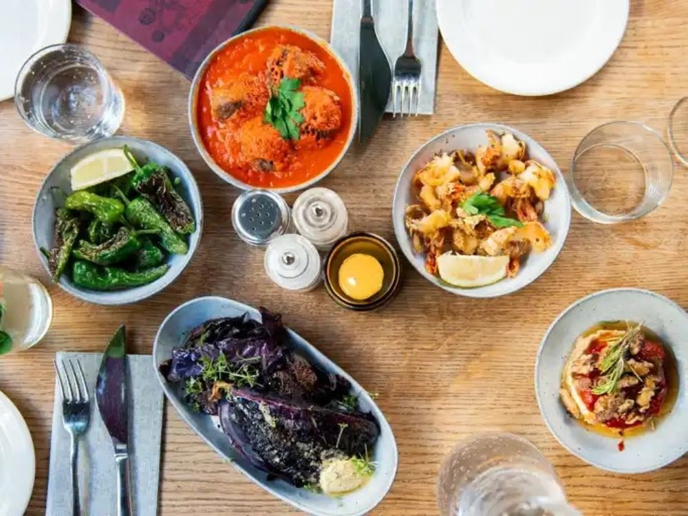 A vibrant spread of dishes including spicy shrimp, roasted vegetables, and dips on a wooden table, with drinks and cutlery nearby.