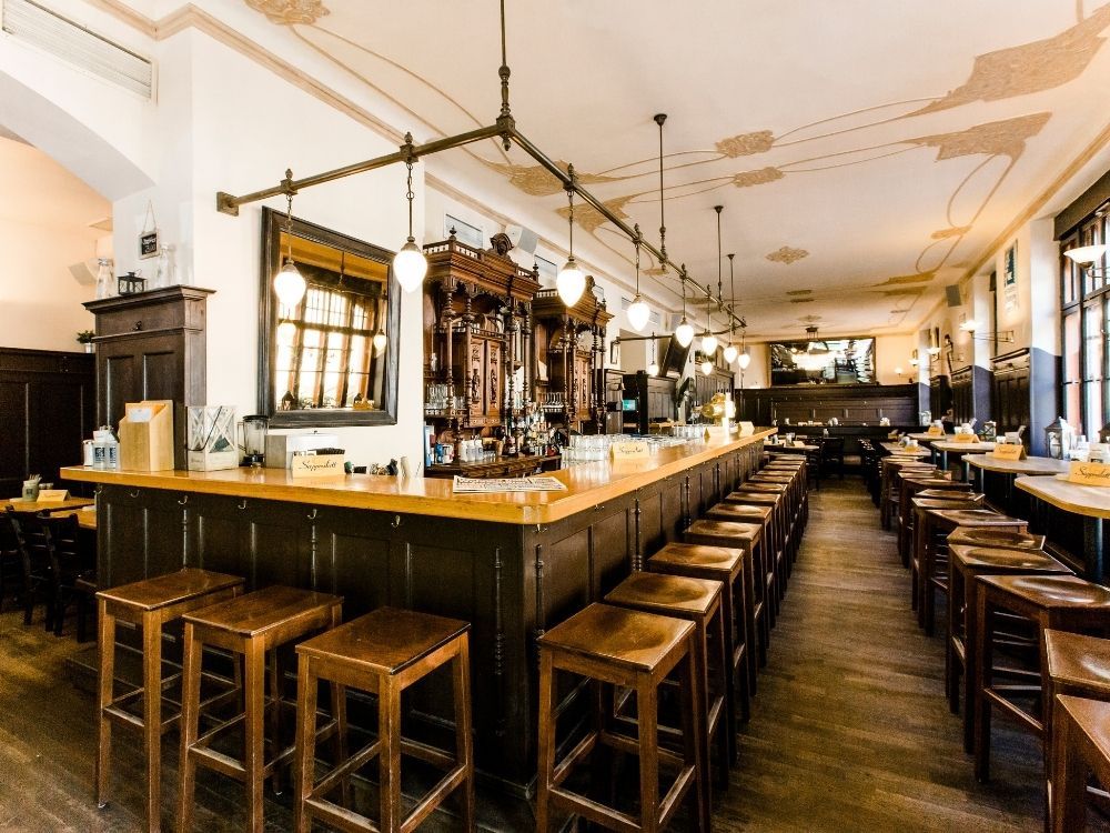 A spacious bar with a long wooden counter, high stools, and elegant decor, featuring ambient lighting and a vintage wooden backbar.