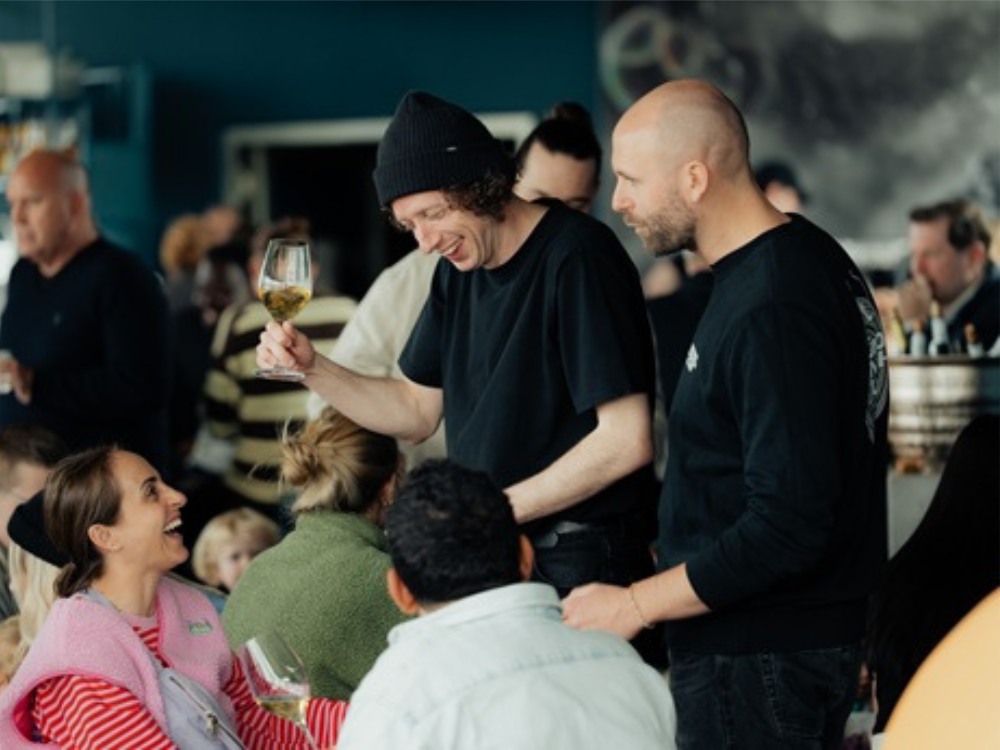 A lively social gathering with people smiling and chatting, some holding wine glasses. The atmosphere is warm and friendly in a dimly lit setting.