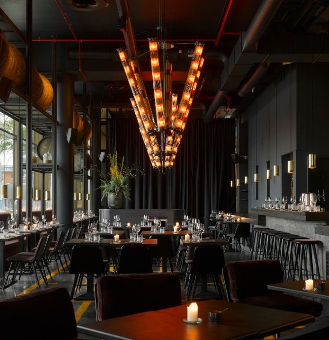 Modern restaurant interior with dim lighting, featuring a striking, large chandelier. Dark decor, tables set with glasses and candles.