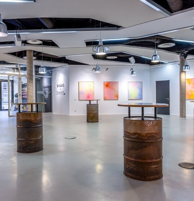 Modern art gallery with industrial decor. Rusted barrels used as tables contrast the sleek gray floor. Abstract paintings with vibrant colors adorn the white walls.