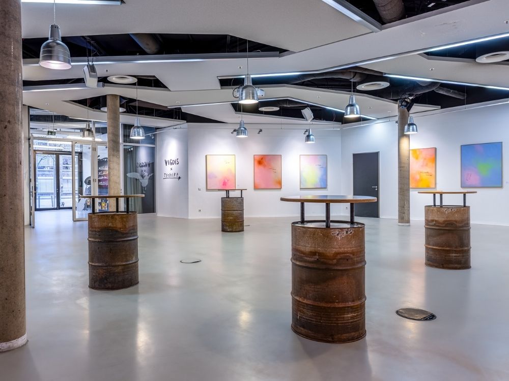 Modern gallery interior with abstract art on white walls and industrial-style tables made from barrels. Bright, open space with ceiling lights.