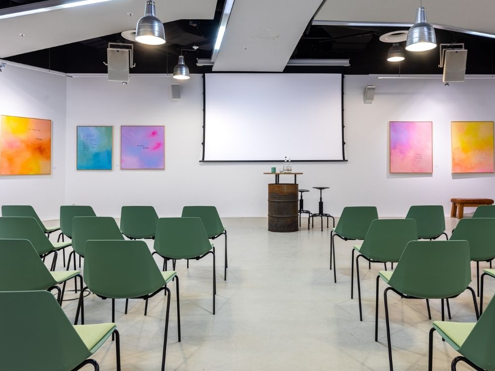 A modern conference room with green chairs arranged facing a blank projection screen and bright abstract paintings adorn the white walls.