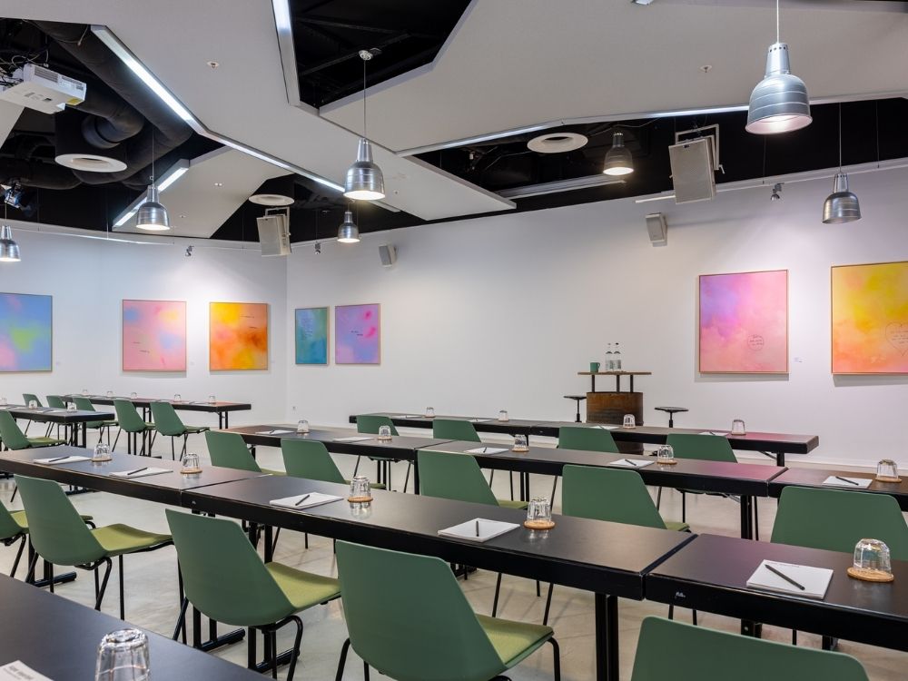 Bright conference room with rows of tables and green chairs, featuring abstract colorful artwork on white walls. Pendant lights hang from the ceiling.