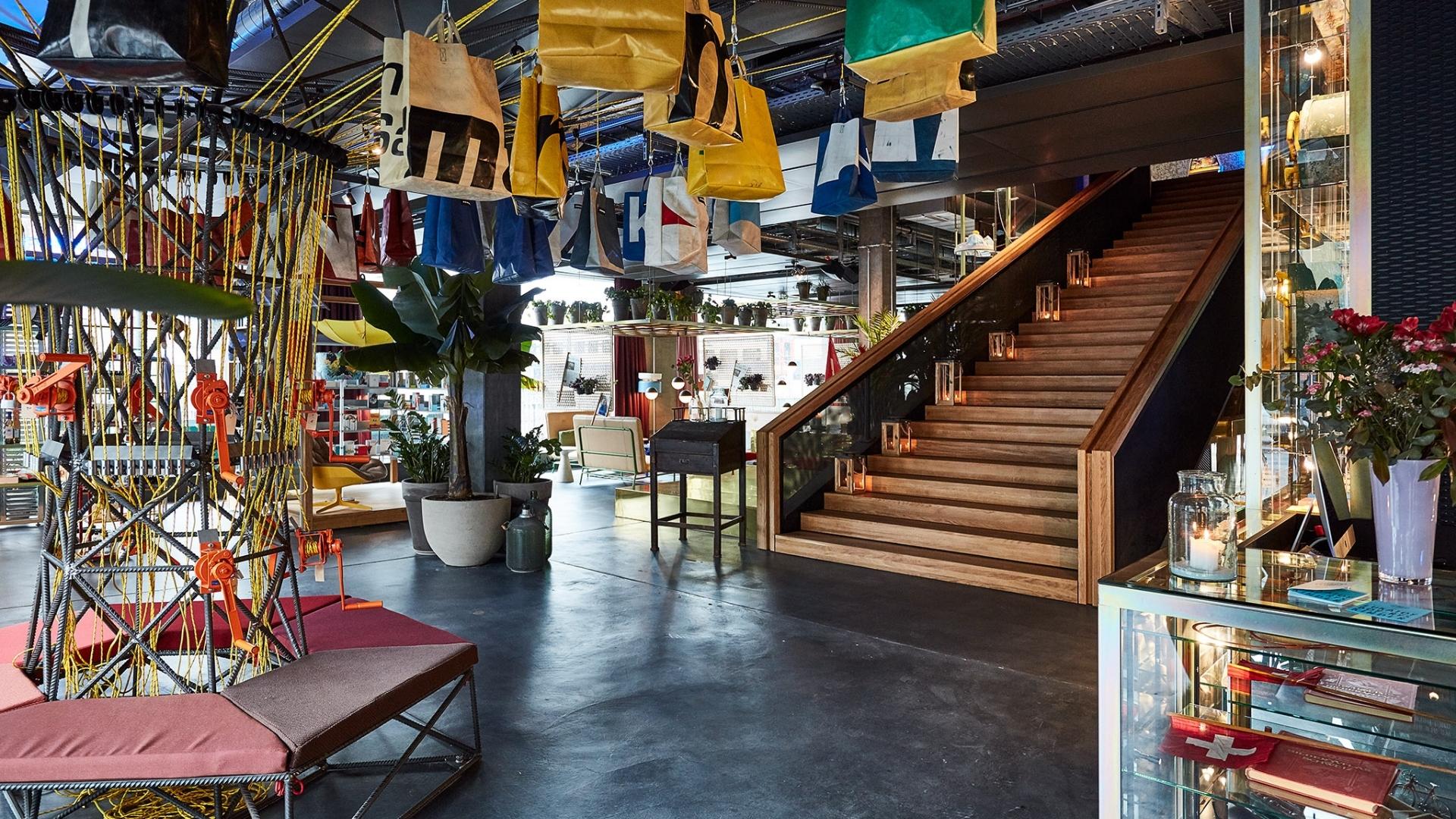 Modern lobby with hanging colorful bags, wooden staircase lined with candles, and a circular seating area. Plants and books add a cosy touch.