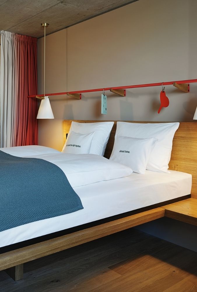 Modern bedroom with a wooden bed featuring white sheets and pillows. A blue blanket covers part of the bed. Two hanging pendant lights and colorful curtains add a cosy atmosphere.