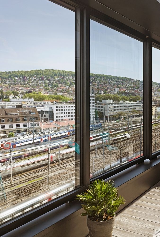 View from a large window showing a bustling train station with multiple tracks and trains. In the foreground is a potted plant. A cityscape and lush green hill are visible in the background.