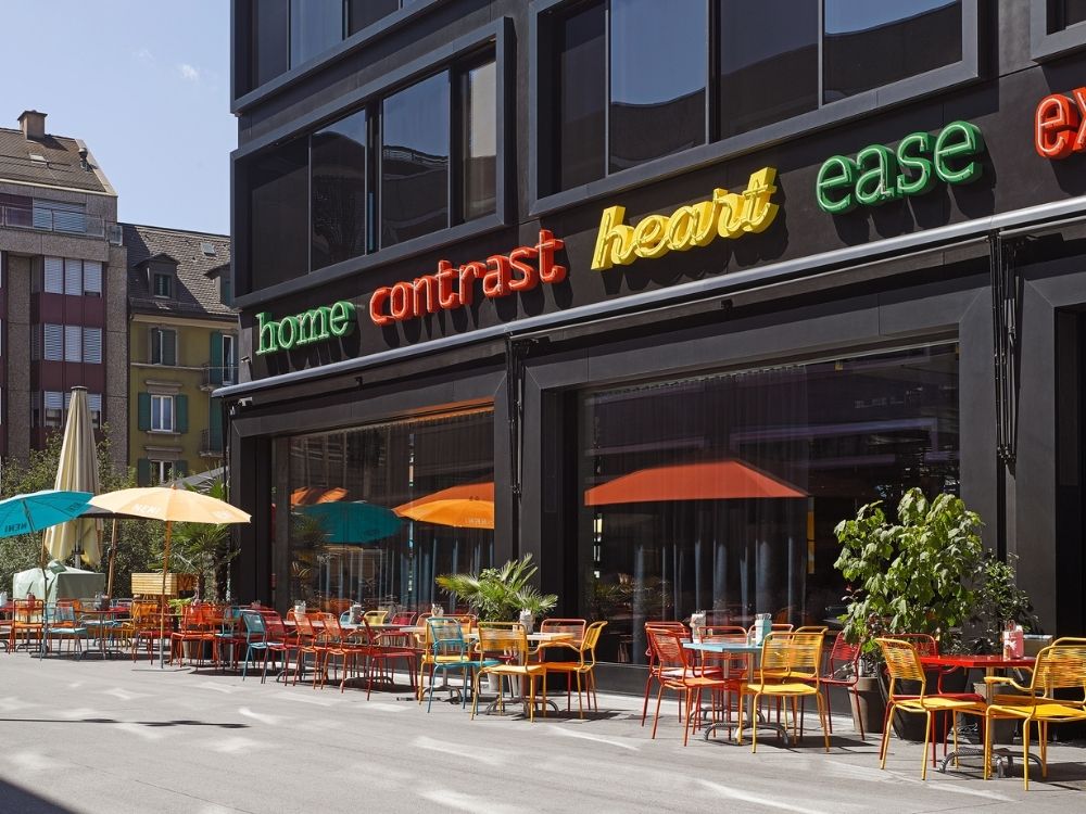 Hotel exterior with vibrant chairs and tables, and bright umbrellas. Neon words 