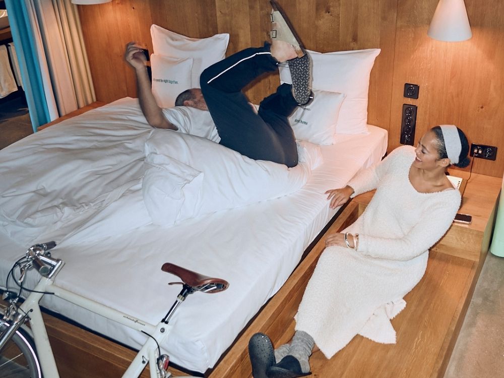 A man in casual clothing playfully tumbles on a bed, while a woman in a cozy white dress and headband sits nearby, smiling warmly. A bicycle is next to the bed, and the room has wooden accents, creating a cozy, lighthearted atmosphere.