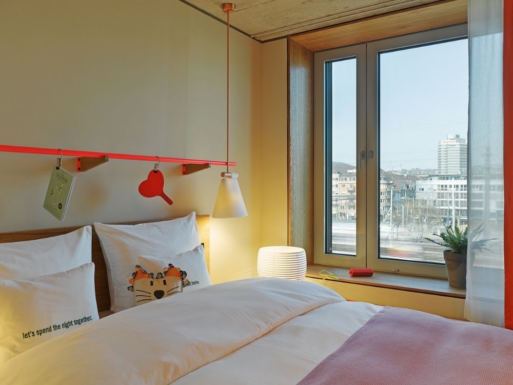 Cosy hotel room with a large window offering a city view. The bed has white and pink bedding, pillows with cute designs, and soft lighting.