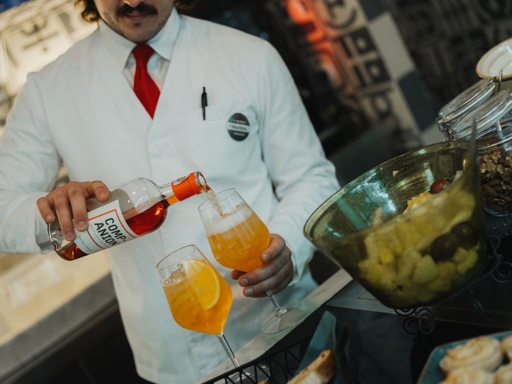 Man in white and red attire pours orange aperitivo into glasses with orange slices. Nearby, a bowl holds mixed fruits. The setting feels lively and festive.