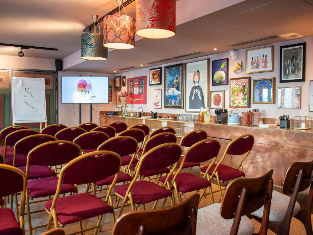 A well-lit conference room with rows of red chairs facing a screen displaying a logo. Artworks and colorful lamps add a creative, inviting atmosphere.