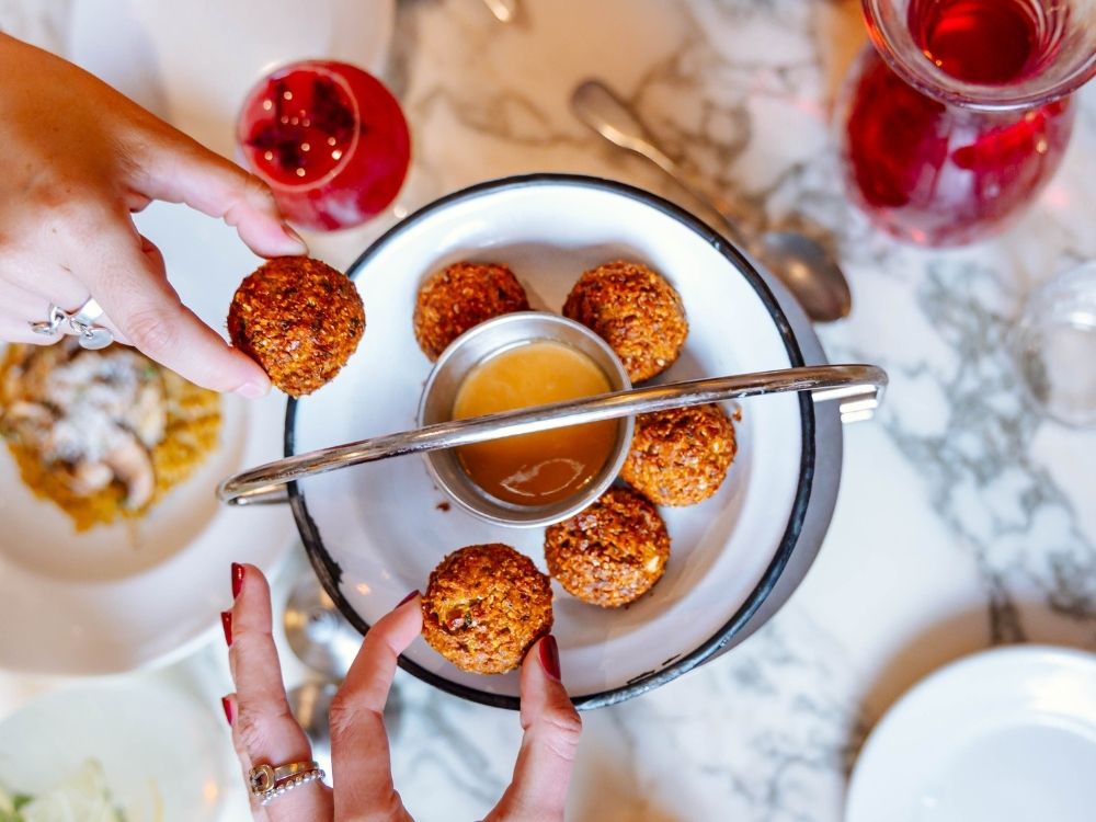 Hands reach for crispy falafel around a dipping sauce on a white plate. The table features red drinks and a dish topped with mushrooms, creating a lively, inviting scene.