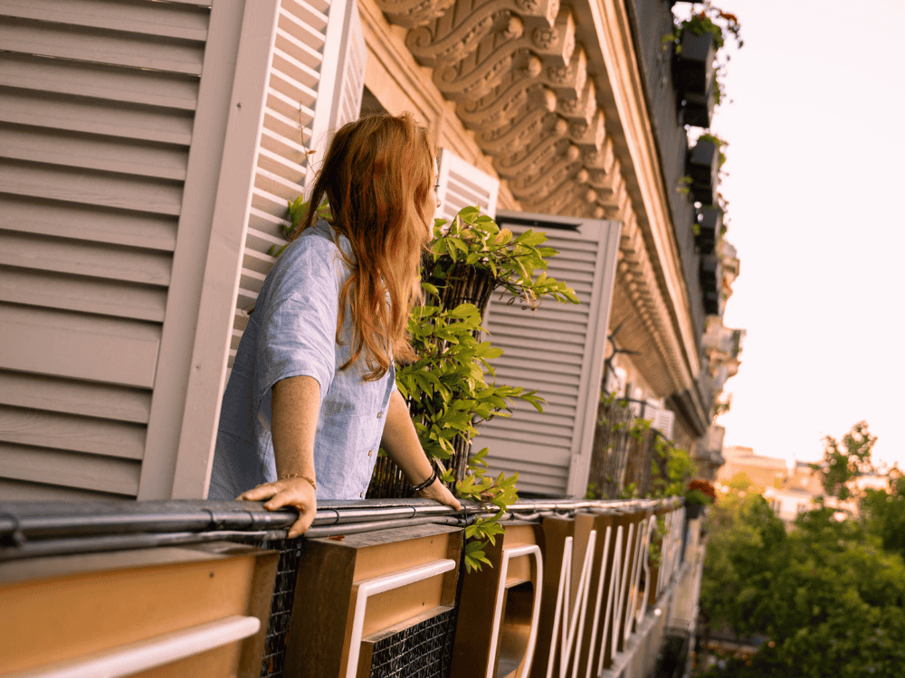 Woman with long hair leans on a balcony railing, gazing towards the sunset. She's surrounded by green plants, creating a peaceful, reflective mood.