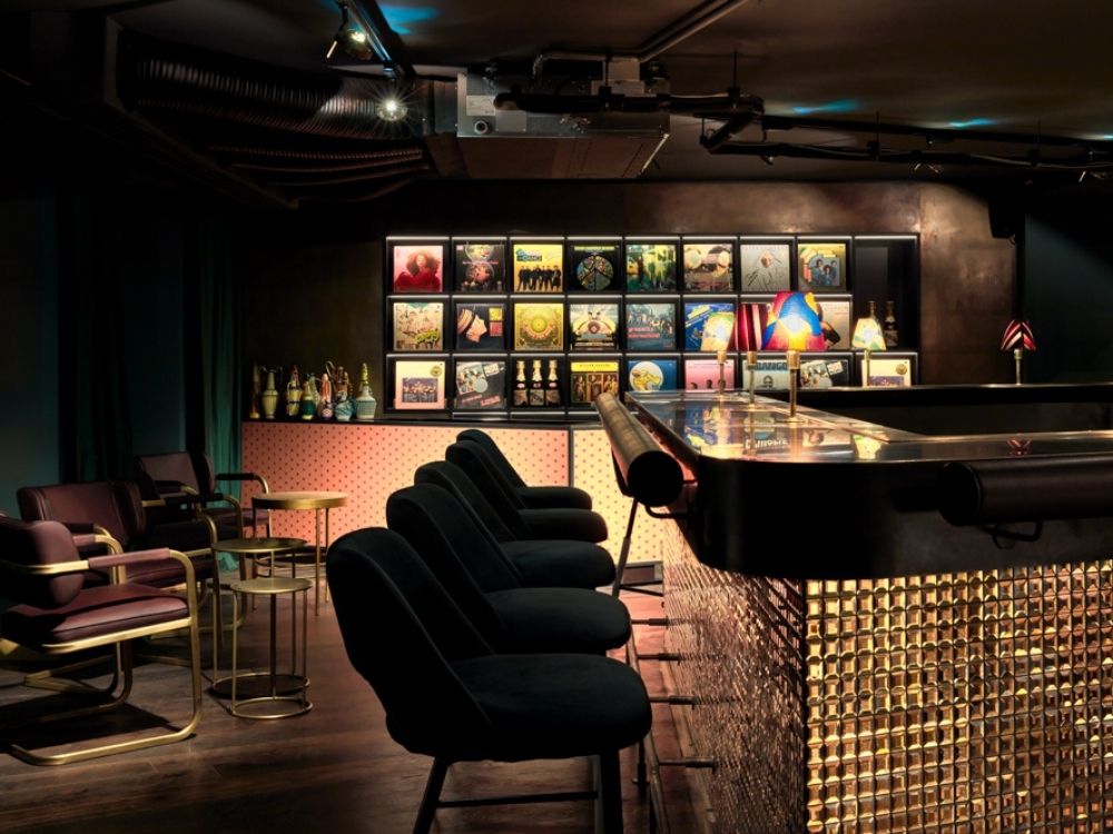 A stylish bar lounge with velvet chairs, illuminated bar, and vintage vinyl record covers on the wall. Warm lighting creates a cosy atmosphere.