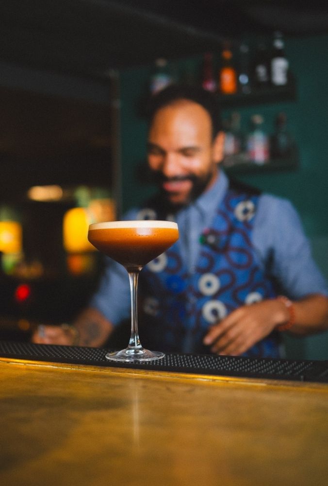 A bartender with a friendly smile stands behind a wooden bar, focusing on an Espresso Martini cocktail in a glass. The setting is dimly lit and cosy.