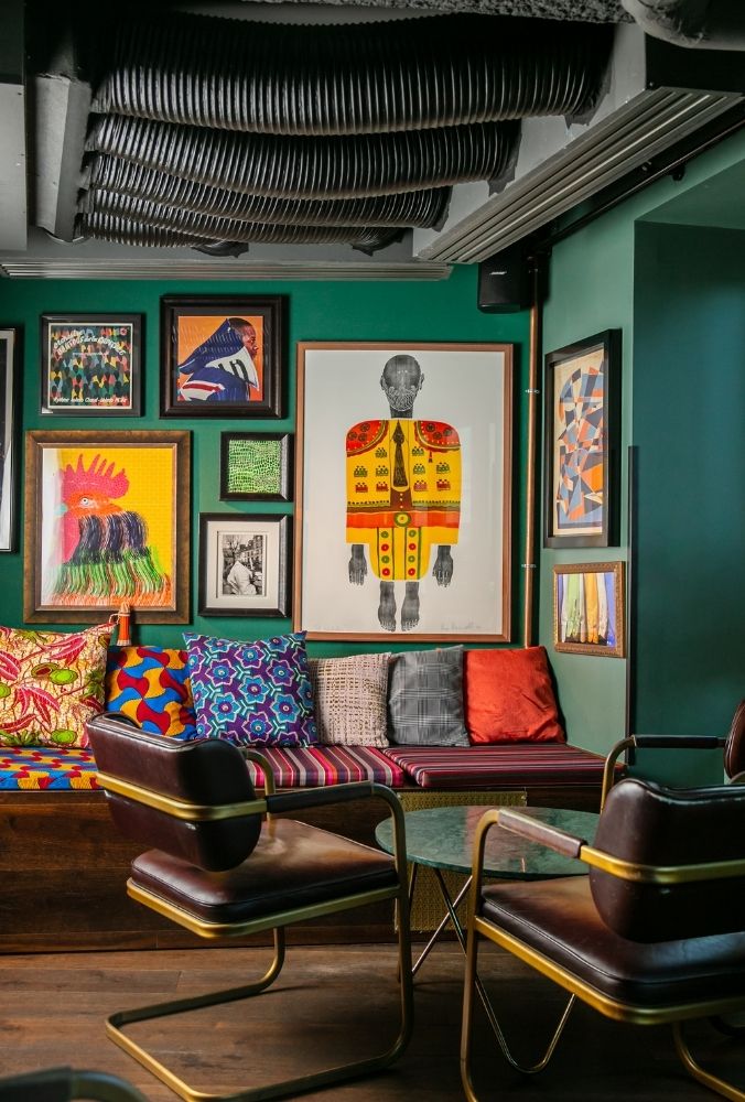 Eclectic room with vibrant art gallery wall, colorful cushions on a bench, mid-century chairs, and industrial ceiling ducts. Creative and cosy ambiance.