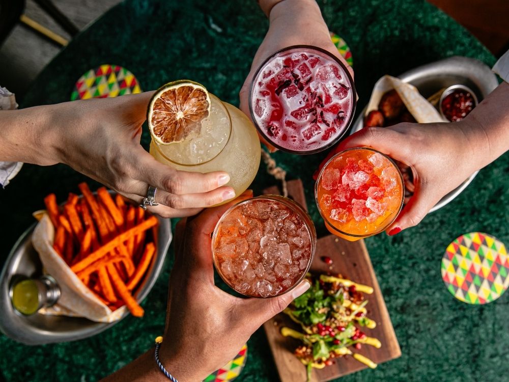 Four hands hold colorful cocktails over a table with sweet potato fries and a vibrant salad, set on a green surface, conveying a joyful, celebratory mood.