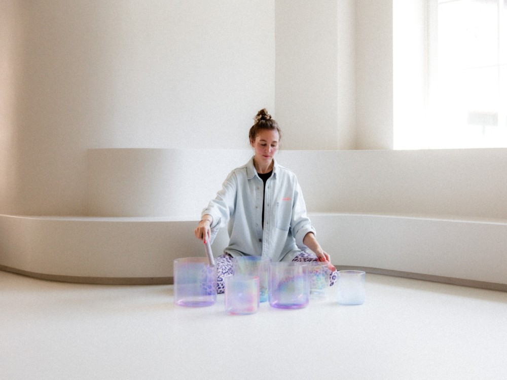 Woman seated in a bright, minimalist room, playing iridescent crystal singing bowls. The serene atmosphere suggests calm and focus.