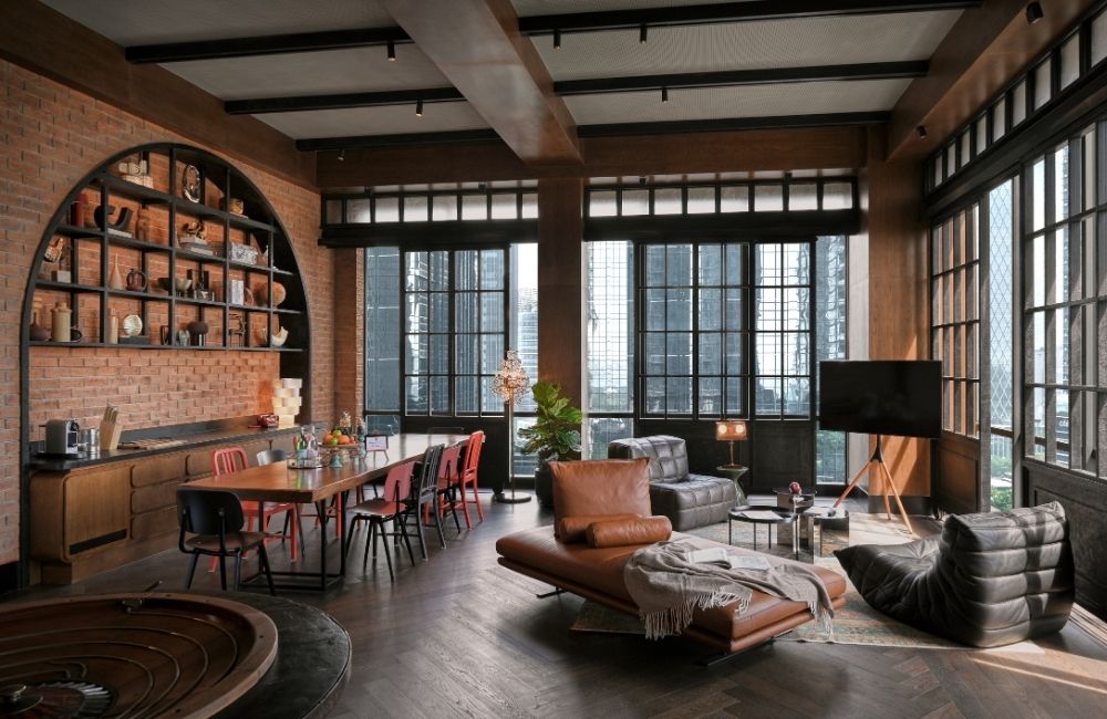 Spacious industrial-style living room with brick walls, large windows, and wooden beams. Features a dining table, leather sofas, and modern decor.