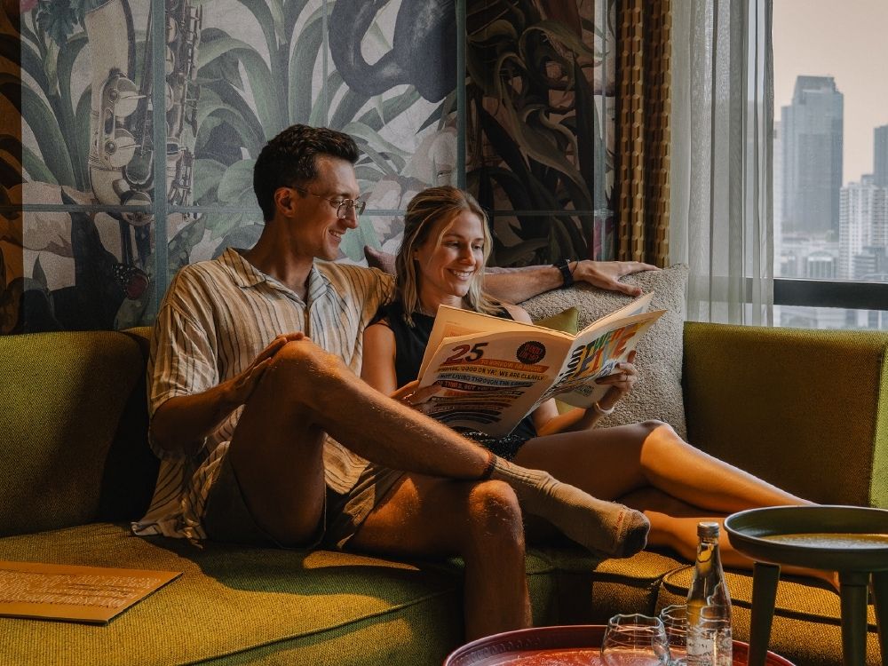 A couple relaxes on a green couch, smiling and reading a magazine with "25" on the cover. A cityscape is visible through a large window beside them.