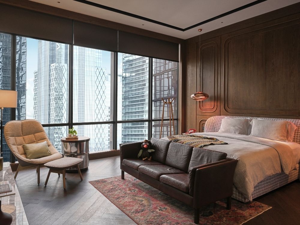 Modern bedroom with wooden walls and large windows revealing a city skyline. Includes a plush bed, leather sofa, armchair, and an inviting, cozy atmosphere.