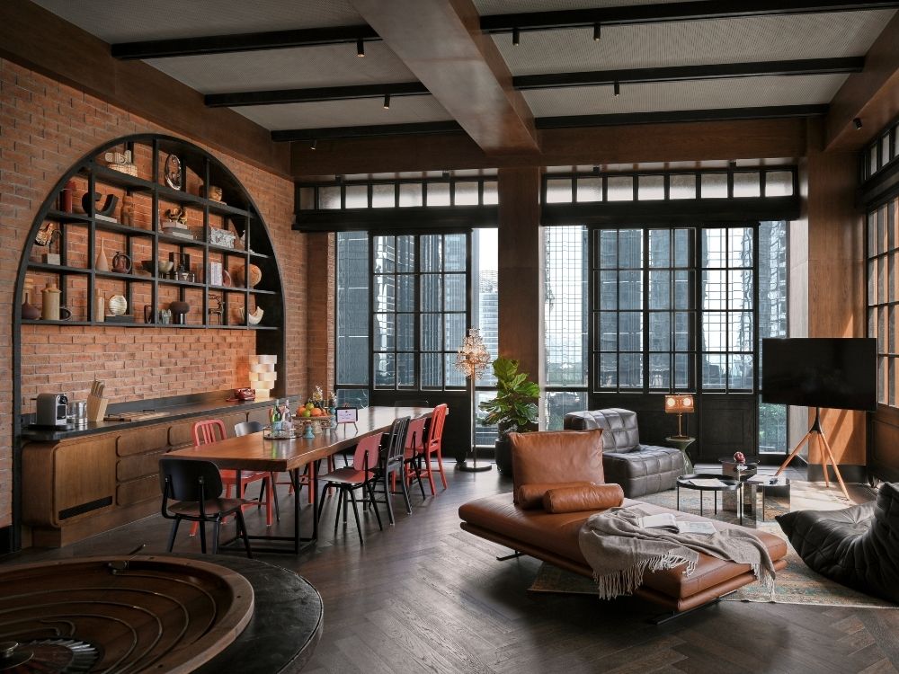 Spacious loft interior with large windows, exposed brick walls, and dark wooden floors. Features a dining table, leather sofa, and modern décor.