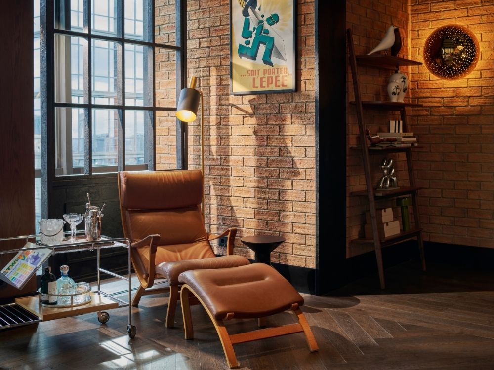 Cosy room with a leather lounge chair and ottoman on dark wood floors. A bar cart and modern bookshelf sit nearby. Warm lighting creates a relaxed ambiance.