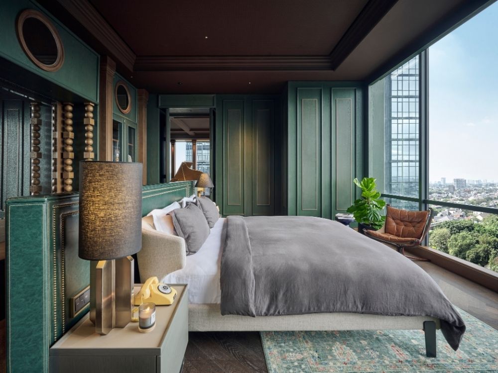 Luxurious bedroom with a large gray bed, green paneled walls, modern lamps, and a cosy chair. Floor-to-ceiling windows reveal a cityscape view.
