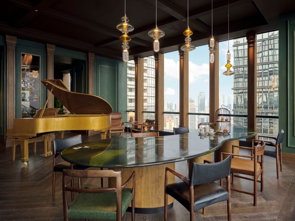 Elegant room with a large green marble table, wooden chairs, and a grand piano. Tall windows showcase a city skyline.