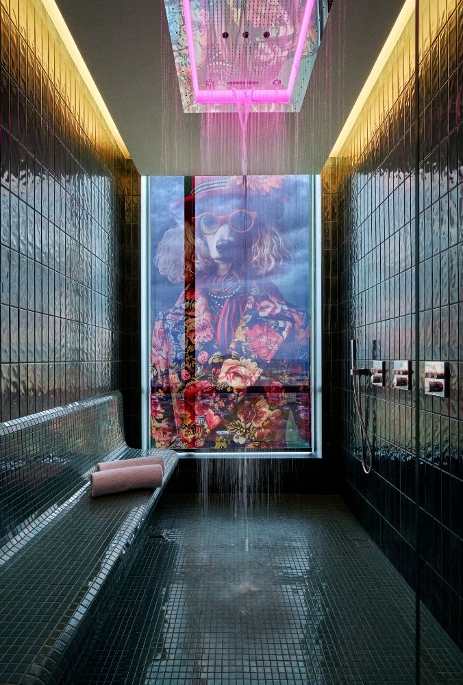 Shower room with green tiles and a ceiling showerhead emitting pink light. A stylized dog portrait with flowers covers the far wall, adding a quirky touch.