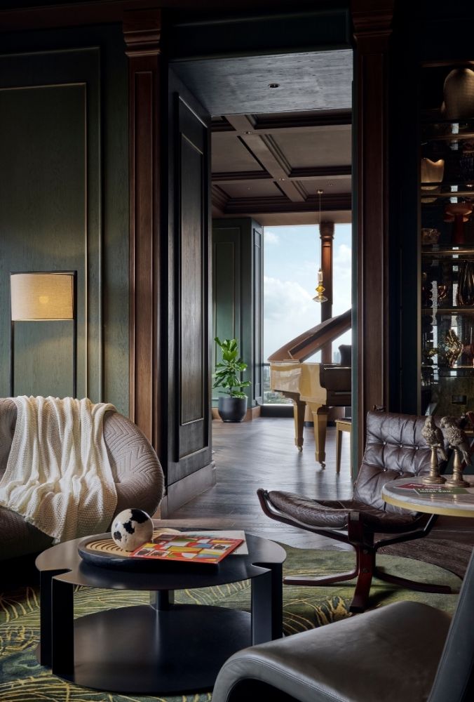 Luxurious living room with dark wood and green walls, featuring a cozy armchair, coffee table, and decor. View into a room with a grand piano and plant.