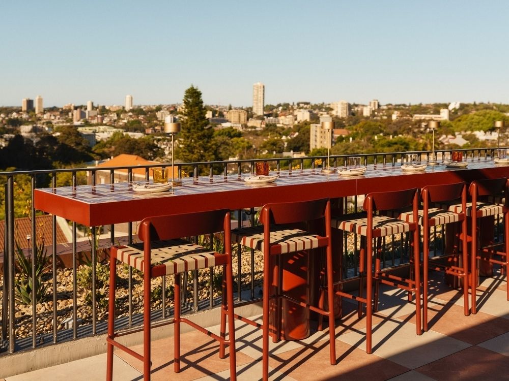 Rooftop patio with a long, red table and matching high chairs, set with plates and glasses. Urban skyline view under clear blue sky.
