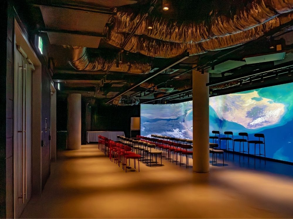 A dimly lit, modern conference room with red chairs, black stools, and a large, vibrant curved screen displaying abstract blue and green visuals.