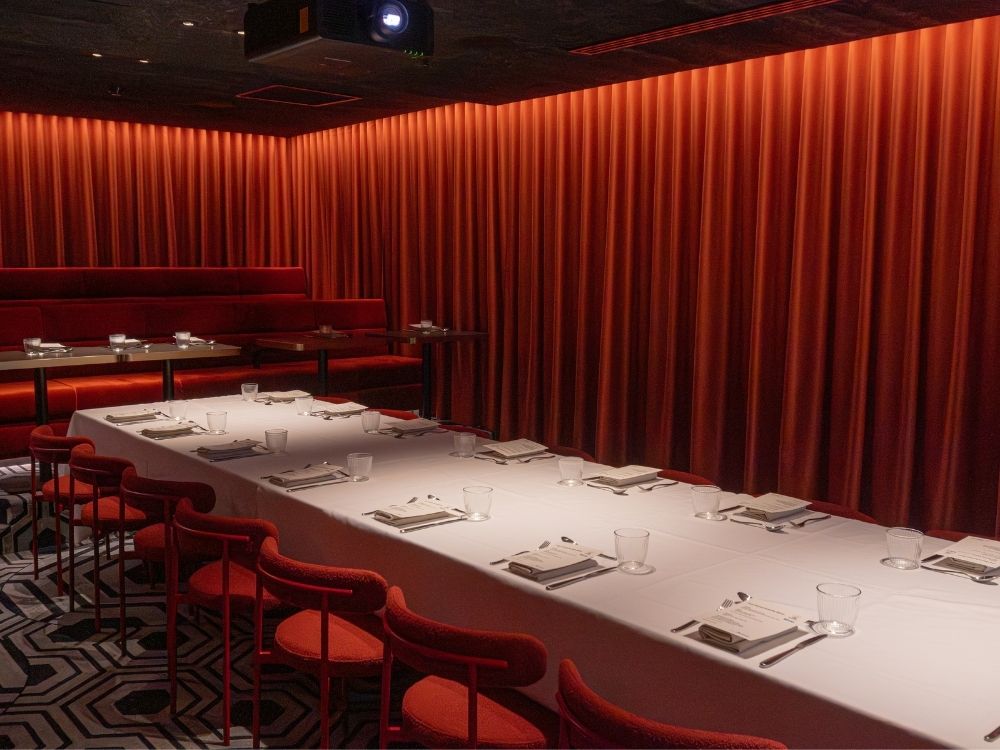 A sophisticated dining room with a long table set for a meal, surrounded by red chairs and illuminated with warm, ambient lighting and red drapes.