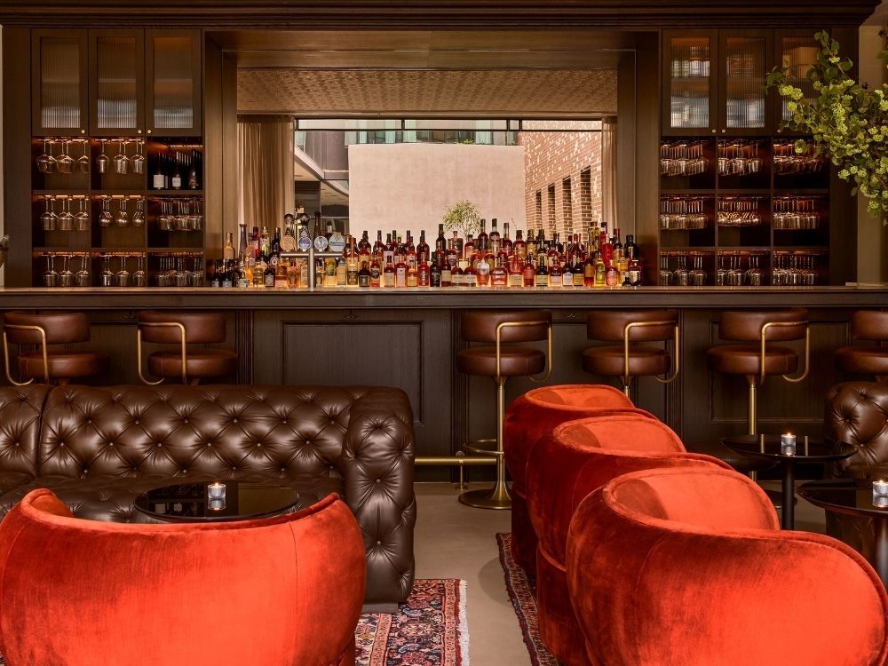 Elegant bar interior with brown leather sofas and vibrant red velvet chairs. A well-stocked bar and warm lighting create a cosy, inviting atmosphere.