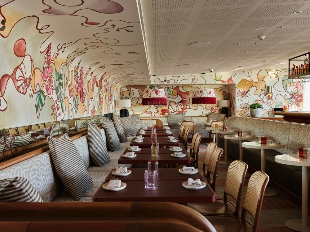 Stylish restaurant interior with colorful abstract wall art, long red tables, cushioned booth seating, and woven chairs. Bright, inviting atmosphere.
