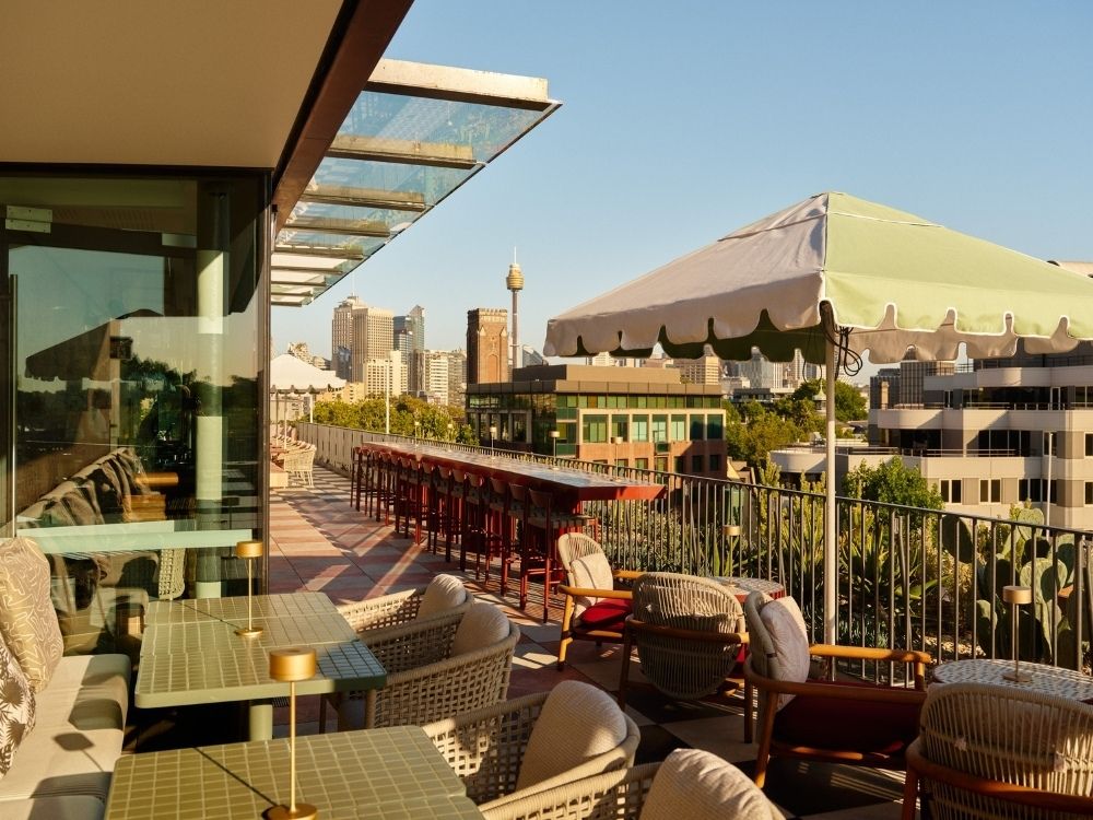 Rooftop patio with cushioned chairs and tables under green-and-white umbrellas, overlooking a city skyline with clear blue skies.