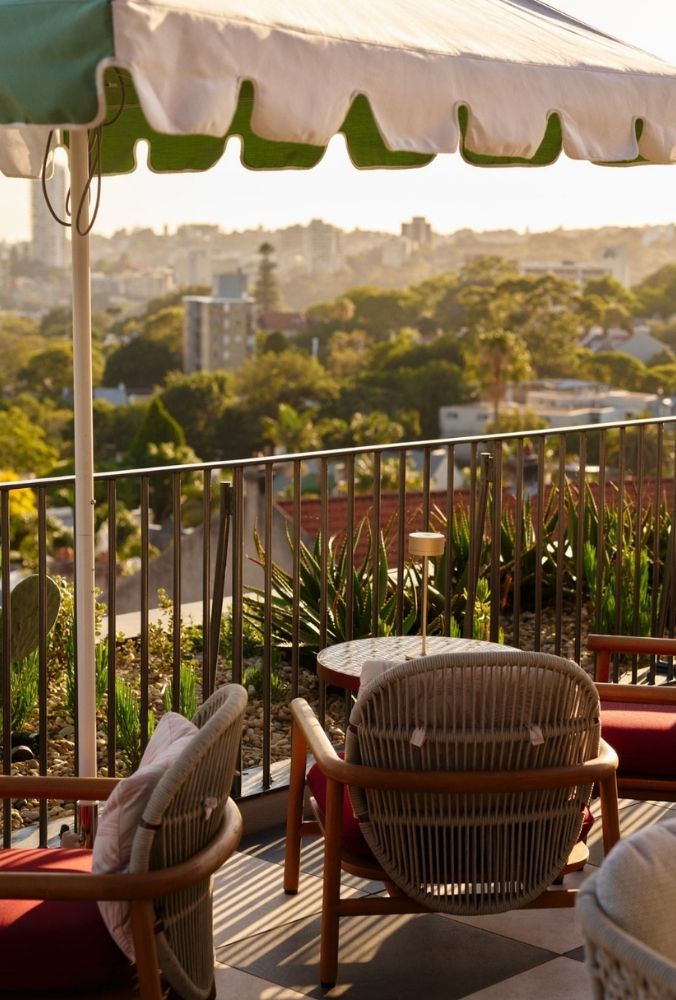 Outdoor terrace with wicker chairs and a round table under a green-and-white striped umbrella. Lush plants and city skyline in the background.