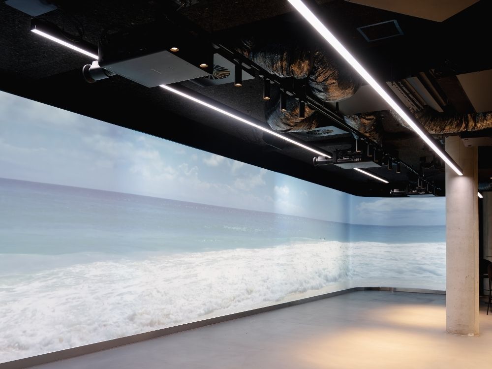 Modern room with a large, wall-sized ocean wave image, evoking a serene beach atmosphere. Overhead lights and exposed ducts add an industrial touch.