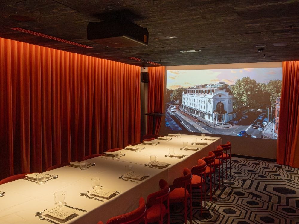 A dimly lit, elegant dining room with a long table set for eight against a backdrop of rich red curtains. A large wall projection shows a city street scene and the 25hours Hotel The Olympia.