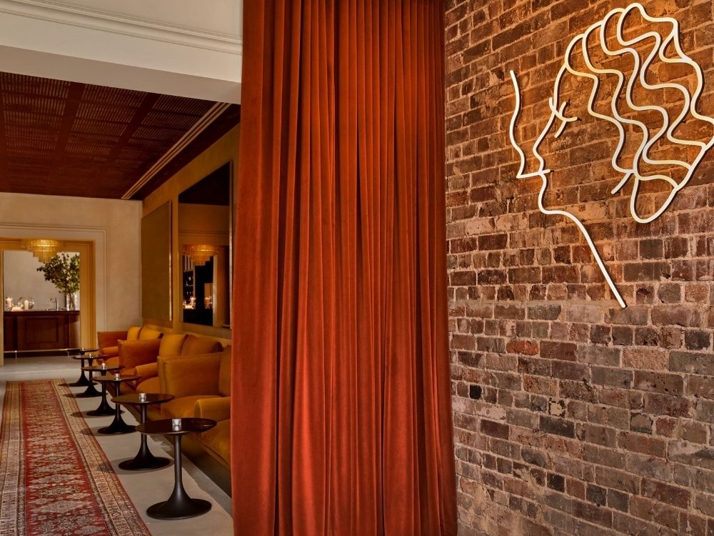 Stylish interior with a warm tone features exposed brick, a neon line art face, rust-colored curtains, gold sofas, and dark side tables.
