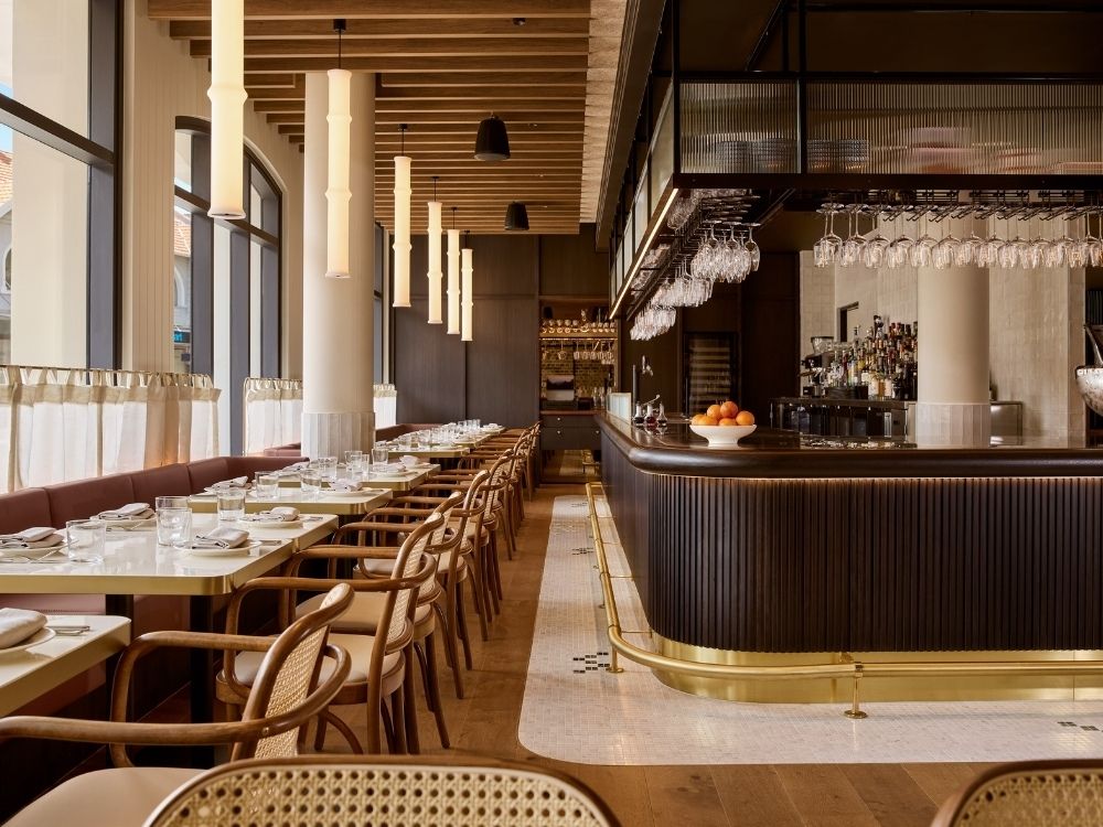 Elegant restaurant interior with wooden floors and ceiling beams, warm lighting, and tables set with white dishes. A sleek bar features hanging glasses and a bowl of oranges.