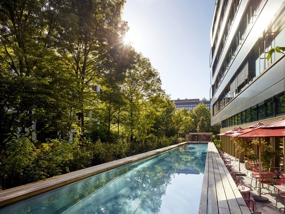 Sunlit outdoor pool beside a modern hotel with red umbrellas. Surrounded by lush green trees, the serene setting conveys tranquility and nature.