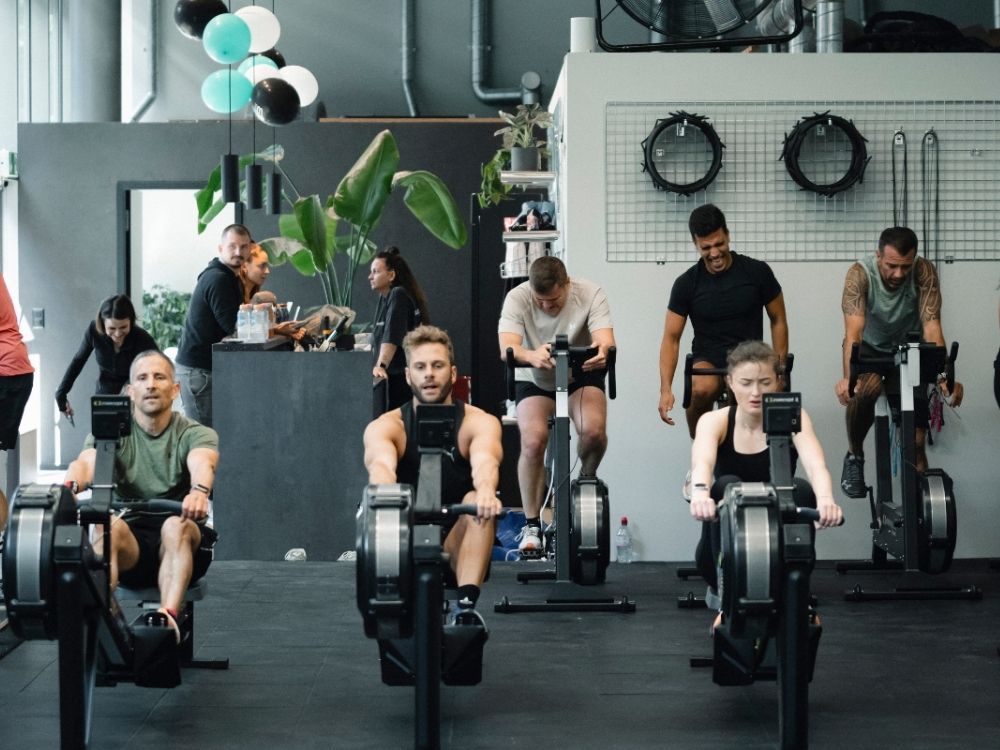 Three people focused and exercising on rowing machines in a gym, with others in the background near a counter. The environment feels busy and energetic.