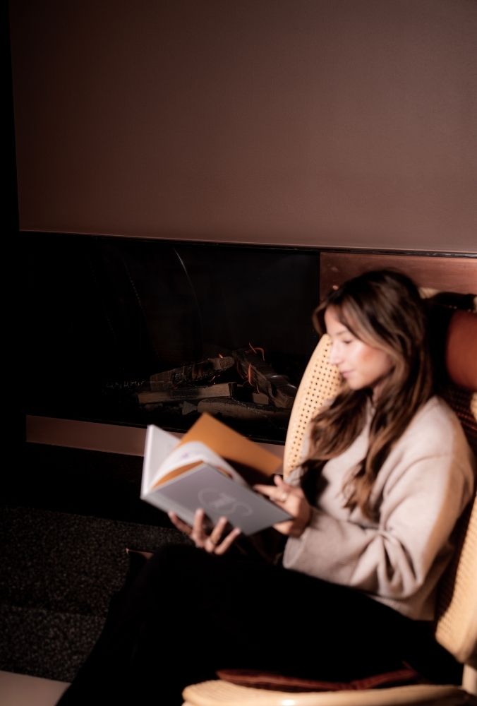 Person sitting in a chair near a fireplace, reading a book, in a cozy, dimly lit room.